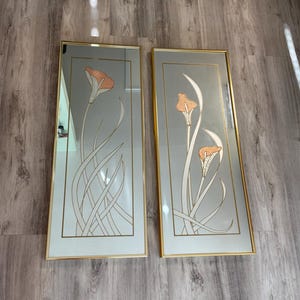 May include: Two rectangular wall mirrors with thin gold frames. Each mirror features a central etched design of calla lily flowers with textured copper-colored petals and long, slender white stems enclosed within a thin gold rectangular border.