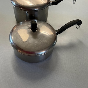 May include: Two stainless steel saucepans with lids and black handles. The pans are different sizes, with one stacked on top of the other. The lids have black knobs and the handles have a loop for hanging.