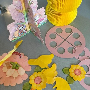 May include: A collection of Easter-themed paper decorations, including a pink and brown tree with bunny illustrations, yellow honeycomb balls, pink circles, and yellow flower cutouts. The decorations are arranged on a gray surface.