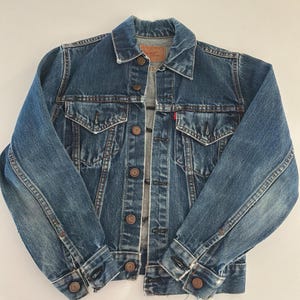 May include: A medium-wash blue denim jacket featuring a classic collar, long sleeves with buttoned cuffs, two chest pockets with button-down flaps, and a front button closure. A small red tab is visible on the left chest pocket seam.