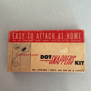 May include: A box of Dot Snappers laundry-proof fasteners. The box is brown with red lettering and a red and white logo. The text on the box reads "Easy to Attach at Home" and "No Sewing - They Go On In A Jiffy!"