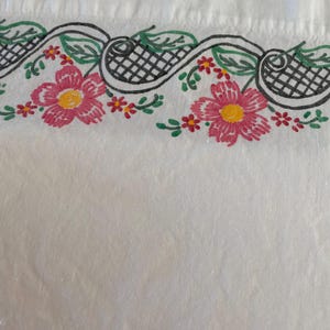 May include: A white fabric textile featuring a decorative embroidered border. The design consists of pink flowers with yellow centers, green leaves, and black cross-hatched patterns connected by flowing black lines on a plain white background.