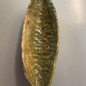 May include: A long, narrow ceramic dish with a textured, green and brown glaze. The dish is shaped like a leaf and has a slightly curved edge.