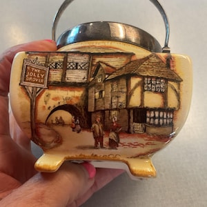 May include: Cream-colored ceramic sugar bowl with a silver metal handle and rim. The bowl features a detailed illustration of a village scene with the text "THE JOLLY DROVER" on a sign. The bowl has three small feet.