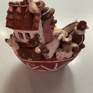 May include: A handcrafted ceramic figurine depicting a boat-shaped ark. The ark is painted in shades of brown and white, with a red roof and a variety of animals and figures inside. The base has a decorative white pattern.