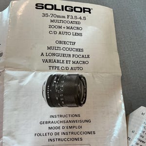 K&ouml;nnte beinhalten: Ein Soligor 35-70mm F3.5-4.5 mehrfach beschichtetes Zoom- und Makro-C/D-Autobjektiv. Das Objektiv ist schwarz mit wei&szlig;en Markierungen und liegt auf wei&szlig;em Papier mit mehrsprachigen Anweisungen. Das Papier hat auch das Soligor-Logo.