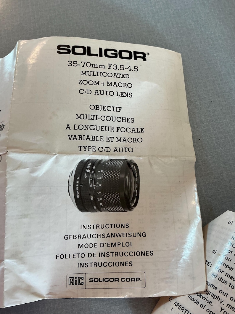 K&ouml;nnte beinhalten: Ein Soligor 35-70mm F3.5-4.5 mehrfach beschichtetes Zoom- und Makro-C/D-Autobjektiv. Das Objektiv ist schwarz mit wei&szlig;en Markierungen und liegt auf wei&szlig;em Papier mit mehrsprachigen Anweisungen. Das Papier hat auch das Soligor-Logo.