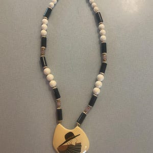 May include: A white and black beaded necklace with a cream-colored pendant featuring a woman wearing a hat and holding a fan.