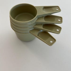 May include: A set of four sage green plastic measuring cups stacked together. Each handle features a small circular hole at the end and is embossed with volume markings including 1/4 cup, 1/3 cup, 1/2 cup, and 2/3 cup in a clear, legible font.