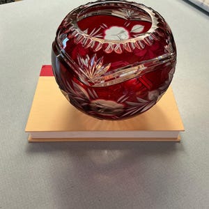 May include: A ruby red, cut crystal bowl with intricate floral and geometric patterns. The bowl is round and sits atop a closed book with a tan cover. The bowl's interior is visible, showcasing a white floral design.