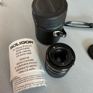 May include: A Soligor 35-70mm F3.5-4.5 zoom lens with a black leather case and a lens cap. The lens has the text "SOLIGOR" and "35-70mm F3.5-4.5 MULTICOATED ZOOM + MACRO C/D AUTO LENS" printed on the packaging.