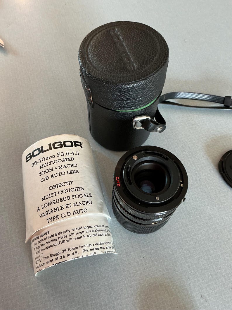 K&ouml;nnte beinhalten: Ein Soligor 35-70mm F3.5-4.5 Zoomobjektiv mit schwarzem Lederetui und Objektivdeckel. Auf dem Objektiv steht "SOLIGOR" und "35-70mm F3.5-4.5 MULTICOATED ZOOM + MACRO C/D AUTO LENS" auf der Verpackung.