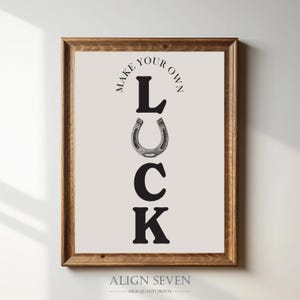 Könnte beinhalten: Ein gerahmter Druck mit den Worten "MAKE YOUR OWN LUCK" in schwarzer Schrift. Der Buchstabe "L" steht über einem Hufeisen, gefolgt von den Buchstaben "C" und "K". Der Rahmen ist dunkelbraun und der Hintergrund hellbeige.