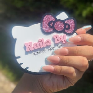 May include: A white, Hello Kitty-shaped sign with a pink glitter bow and the words "Nails By" in pink lettering. The sign also includes the Instagram handle "@pretty.nailz_.bar". The image shows a hand with manicured nails.
