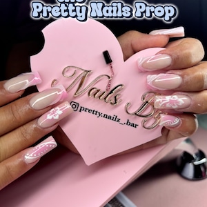May include: Pink and white long square shaped acrylic nails with a white floral design. The nails are displayed on a pink heart-shaped sign with the text "Nails By @pretty.nailz_bar".
