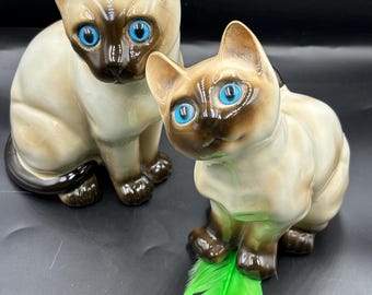 May include: Two ceramic Siamese cat figurines with blue eyes. The larger cat is sitting with its tail curled around its legs. The smaller cat is sitting with its paws tucked under its body. Both cats are painted in shades of brown and white.