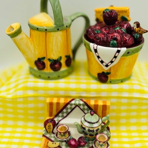 May include: A decorative ceramic kitchenware set with an apple theme. The set includes a watering can, a bucket filled with apples, and a miniature tea set. The pieces are painted yellow, green, and red, with apple and leaf details.