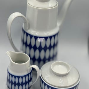 May include: A white ceramic coffee pot, creamer and sugar bowl set with a blue and white geometric pattern. The set features a lidded coffee pot with a spout, a small creamer jug with a handle, and a lidded sugar bowl.