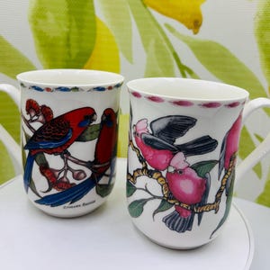 May include: Two white ceramic mugs with bird illustrations. One mug features red and blue parrots on a branch, with the text "Crimson Rosella." The other mug displays pink and grey birds. Both mugs have a white handle and a decorative rim.