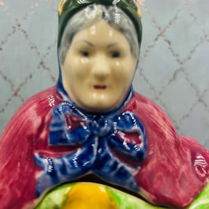 May include: A ceramic figurine of an elderly person wearing a dark green bonnet with yellow floral accents, a bright red shawl, and a blue necktie. The figure has painted facial features, grey hair, and a glossy finish on the glazed ceramic surface.