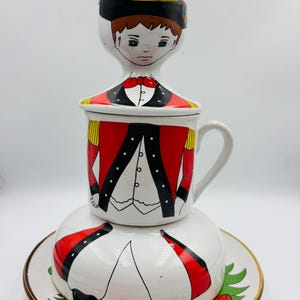 May include: A white ceramic teacup, saucer, and egg cup set with a whimsical design of a boy in a red and black uniform. The boy's face is painted on the egg cup, his torso on the teacup, and his legs on the saucer.