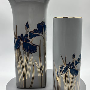 May include: Two grey ceramic vases with gold trim and floral designs. The vases feature blue iris flowers and gold accents on a grey background. The taller vase has a flared top, the shorter one is cylindrical.