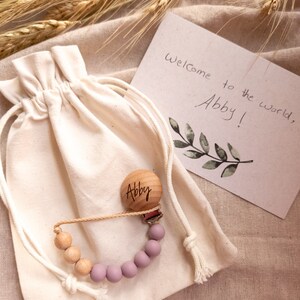 May include: A personalized wooden baby teether with a purple silicone bead chain, packaged in a white drawstring bag. The bag is sitting on a beige fabric surface with wheat stalks. The wooden teether is engraved with the name "Abby". A card with the text "Welcome to the world, Abby!" is visible in the background.