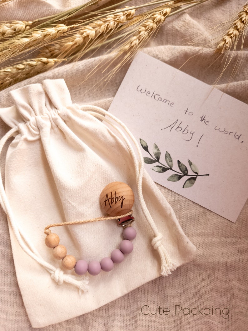 May include: A personalized wooden baby teether with a purple silicone bead chain, packaged in a white drawstring bag. The bag is sitting on a beige fabric surface with wheat stalks. The wooden teether is engraved with the name "Abby". A card with the text "Welcome to the world, Abby!" is visible in the background.