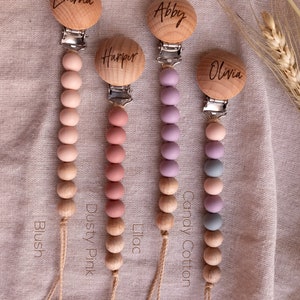 May include: Four wooden baby teethers with personalized names: Emma, Abby, Harper, and Olivia. Each teether has a string of silicone beads in different colors: blush, dusty pink, lilac, and candy cotton.