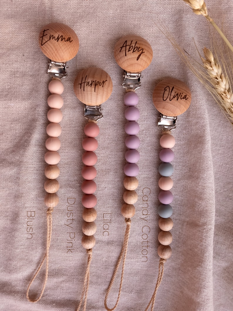 May include: Four wooden baby teethers with personalized names: Emma, Abby, Harper, and Olivia. Each teether has a string of silicone beads in different colors: blush, dusty pink, lilac, and candy cotton.