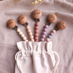 May include: Set of five wooden baby teethers with personalized names: Hudson, Emma, Harper, Abby, and Olivia. Each teether has a silicone beaded chain in a different color: beige, pink, light pink, purple, and blue. The teethers are displayed in a white drawstring bag.