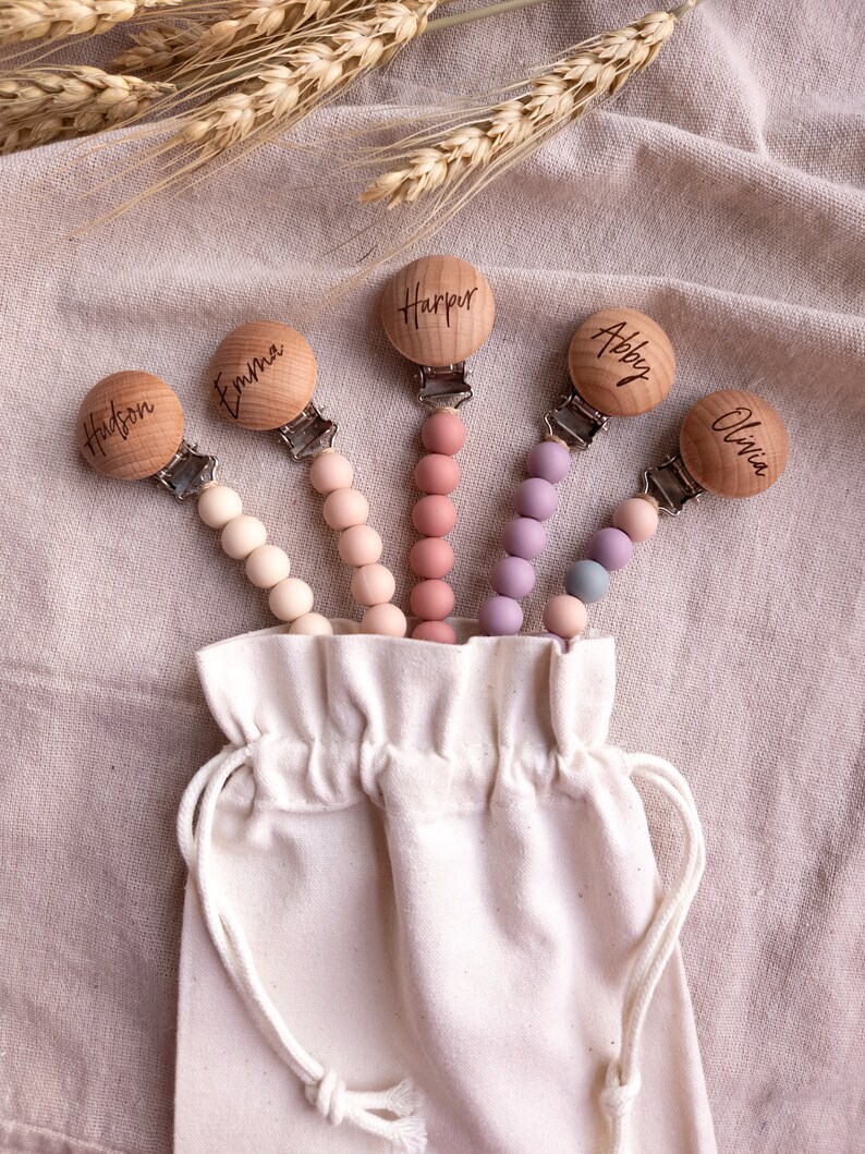 May include: Set of five wooden baby teethers with personalized names: Hudson, Emma, Harper, Abby, and Olivia. Each teether has a silicone beaded chain in a different color: beige, pink, light pink, purple, and blue. The teethers are displayed in a white drawstring bag.