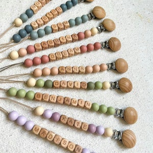 May include: A set of ten personalized wooden bead necklaces with silicone beads in various colors. Each necklace features a different name spelled out in wooden blocks. The necklaces are attached to wooden clips.