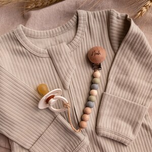 May include: A beige ribbed baby onesie with a wooden pacifier clip attached. The clip has a wooden bead with the name "Noah" engraved on it. The pacifier clip has a variety of colored wooden beads and silicone beads.