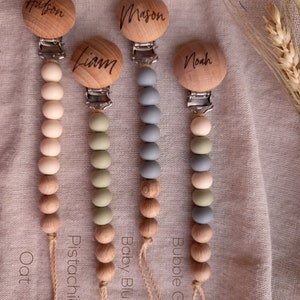 May include: Four wooden baby teethers with different colored silicone beads. The teethers are labeled "Oat", "Pistachio", "Baby Blue", and "Bubble Gum". Each teether has a wooden clip with a name engraved on it: "Hudson", "Liam", "Mason", and "Noah".