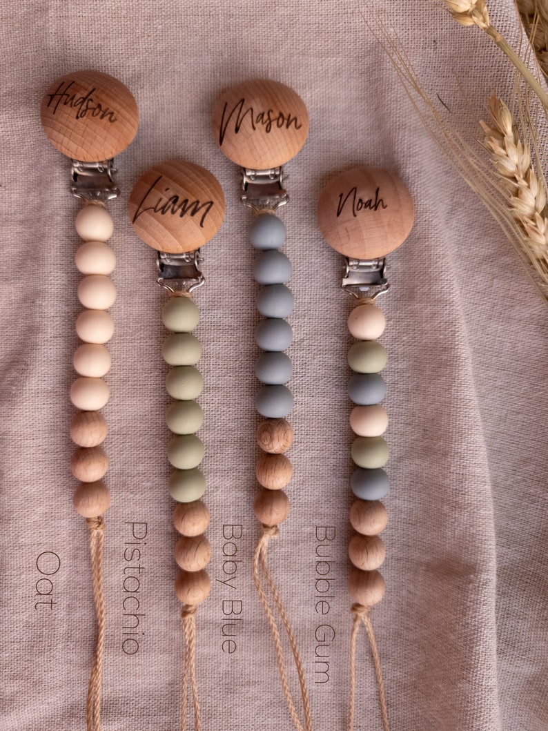 May include: Four wooden baby teethers with different colored silicone beads. The teethers are labeled "Oat", "Pistachio", "Baby Blue", and "Bubble Gum". Each teether has a wooden clip with a name engraved on it: "Hudson", "Liam", "Mason", and "Noah".