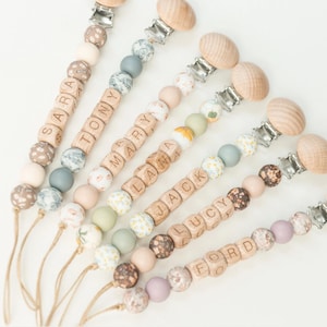 May include: A set of seven personalized wooden baby teethers with different names, including "Sara", "Tony", "Mary", "Liam", "Jack", "Lucy", and "Ford". Each teether features a wooden bead with the name engraved on it, along with a variety of colorful beads and a wooden clip.