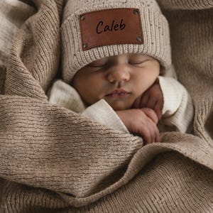 May include: A sleeping baby wrapped in a soft, brown blanket and wearing a beige knit cap with the name "Caleb" on a leather patch. The baby's face is peaceful, and the overall image evokes a sense of warmth and comfort.