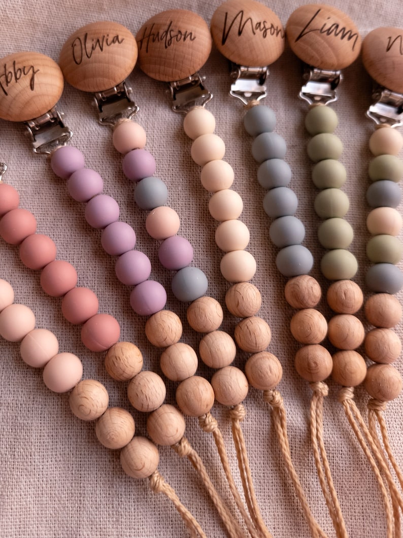 May include: Six wooden teethers with personalized names, each with a different color silicone bead strand. The names are Olivia, Hudson, Mason, Liam, Bobby, and a fifth name that is not visible. The teethers are attached to a natural twine string.