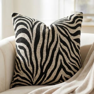 Puede incluir: Cojín decorativo cuadrado con un estampado de rayas de cebra en negro y beige. El cojín está hecho de una tela texturizada y está colocado en un sofá blanco con una manta beige.