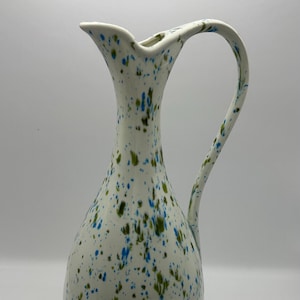 May include: A white ceramic pitcher with a splatter design in shades of blue and green. The pitcher has a wide mouth and a curved handle.