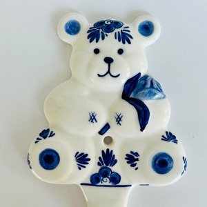 May include: A white ceramic teddy bear figurine with blue and white Delftware designs. The bear is holding a blue flower and has a hole in the center of its chest.