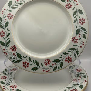 May include: Three white ceramic dinner plates with a gold rim and a green and red floral design. The design features holly leaves, red berries, and poinsettia flowers.