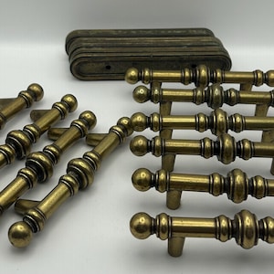 May include: A collection of antique brass cabinet pulls and backplates. The pulls have a detailed, ornate design with a warm, aged finish. The backplates are rectangular with rounded edges. Ideal for vintage furniture restoration.