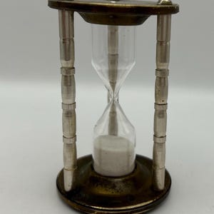 May include: A small, antique hourglass with a brass frame and silver accents. The hourglass is filled with white sand and has a clear glass bulb.