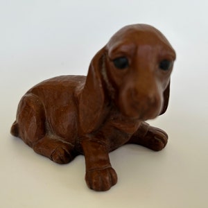 May include: A small, brown, ceramic figurine of a dachshund puppy sitting with its head down. The figurine has a realistic appearance with detailed features, including a long snout, floppy ears, and a curled tail.