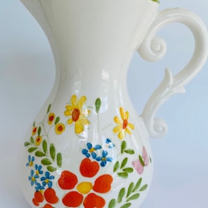 May include: White ceramic pitcher with a green rim and a floral design. The flowers are yellow, orange, red, blue, and pink. There is a butterfly on the side of the pitcher.