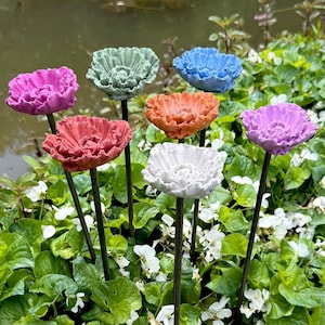May include: A collection of colorful, decorative flower garden stakes. The stakes feature sculpted flower heads in various colors, including pink, green, blue, orange, and white. They are set amidst lush green foliage and small white flowers, with a pond in the background.