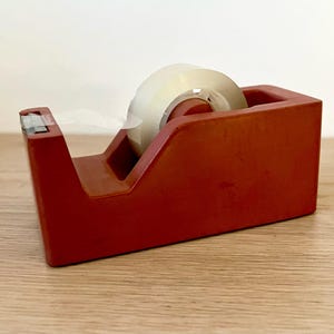 May include: A rectangular red tape dispenser with a weighted base and a serrated metal cutting edge at the front. A roll of clear adhesive tape is mounted on the internal spindle, with the end of the tape pulled forward toward the metal blade.