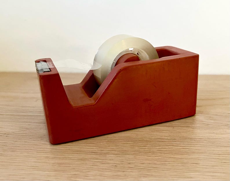 May include: A rectangular red tape dispenser with a weighted base and a serrated metal cutting edge at the front. A roll of clear adhesive tape is mounted on the internal spindle, with the end of the tape pulled forward toward the metal blade.
